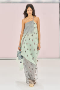 Model walking a runway in a pastel mint-green and gray layered saree with floral embroidery, holding a small beaded clutch.