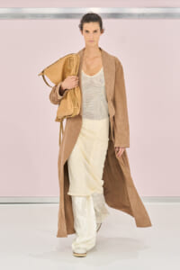 Model walks the runway in a long beige-brown coat over a cream layered outfit, carrying a tan leather bag over her shoulder.