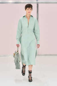 Model walking the runway in a mint green midi shirt dress with an open placket, carrying a matching fringe handbag.