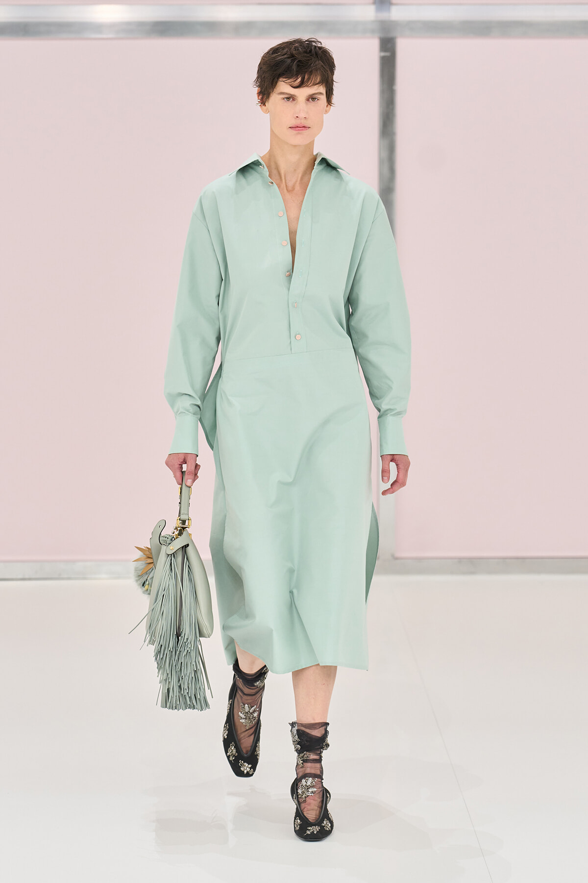 Model walking the runway in a mint green midi shirt dress with an open placket, carrying a matching fringe handbag.