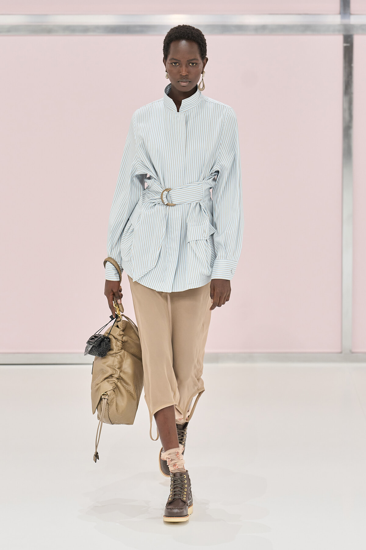 Model walking a runway in a light blue pinstriped blouse cinched with a wide belt over beige cropped pants and brown rugged boots, carrying a tan bag with a small black pouch.