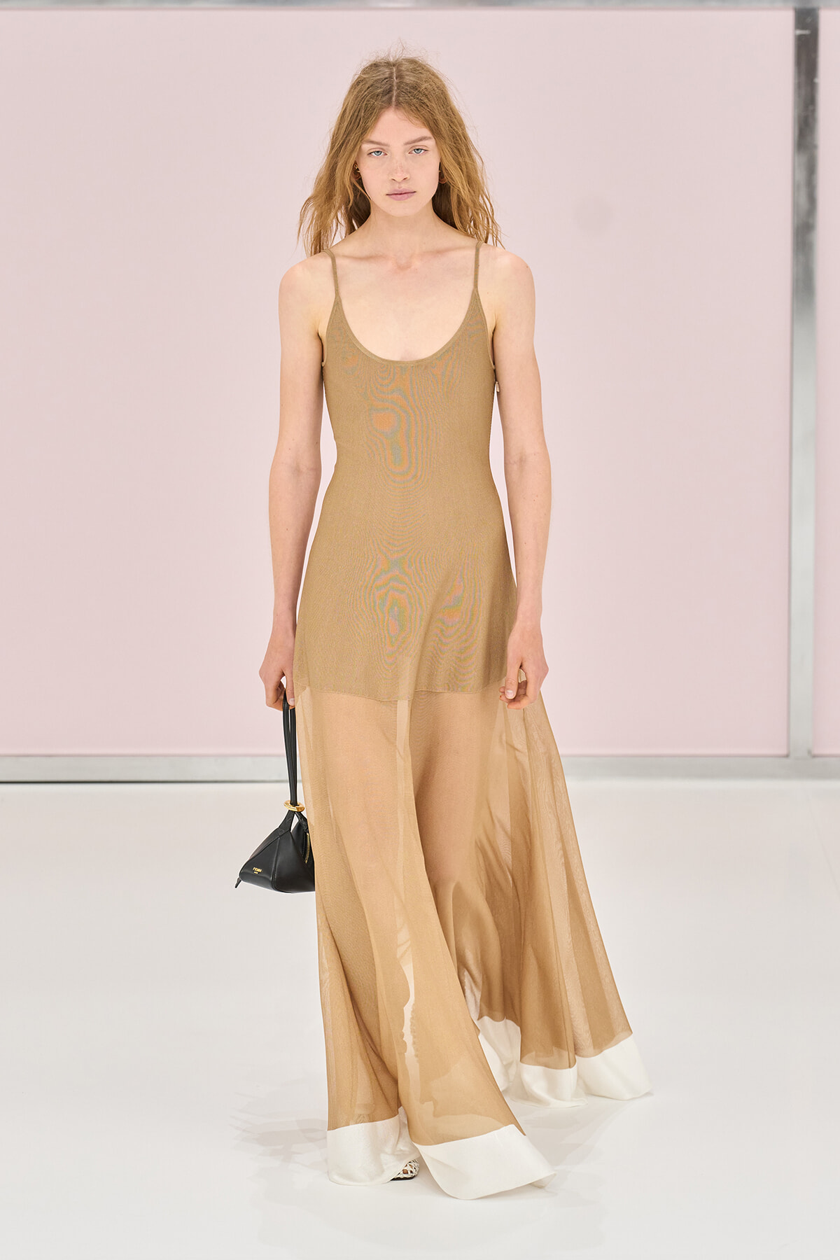 Model walking the runway in a beige sleeveless knit dress with a sheer overlay, a long, flowing skirt with a white hem, carrying a small black handbag.