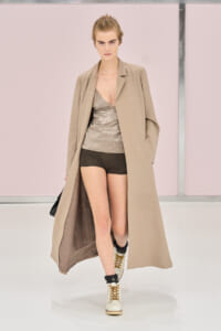 Model walking the runway in a long beige coat over a satin beige top and black shorts, with white lace-up boots.