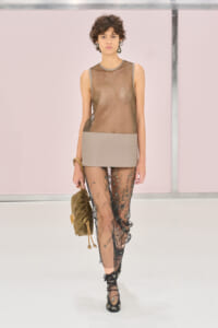 Model on a runway wearing a taupe mesh sleeveless top with a beige panel, sheer embroidered pants, and an olive handbag