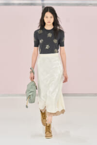 Model walking a runway in a dark floral short-sleeve top and pale satin midi skirt, tan lace-up boots, and a mint green bag.