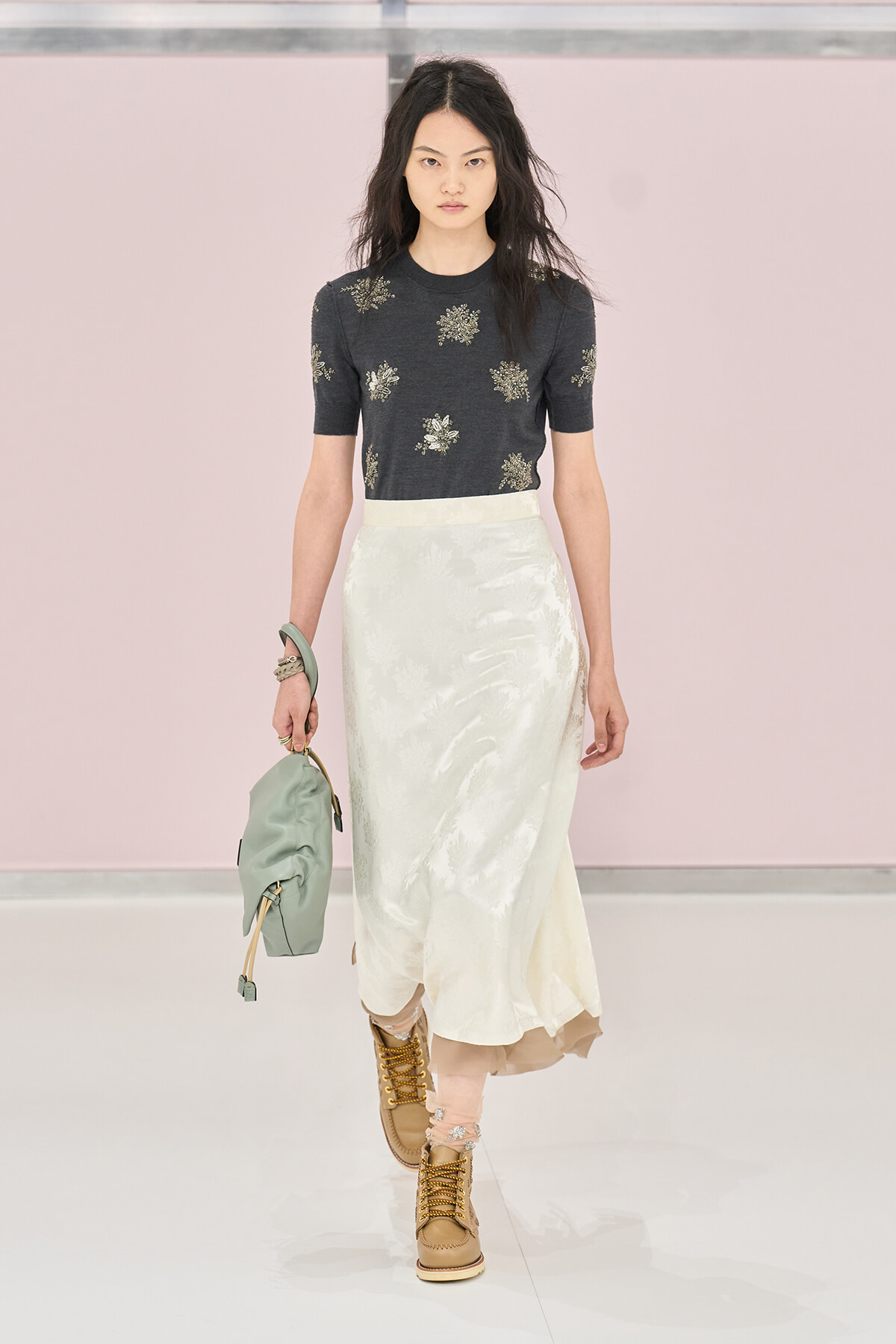 Model walking a runway in a dark floral short-sleeve top and pale satin midi skirt, tan lace-up boots, and a mint green bag.