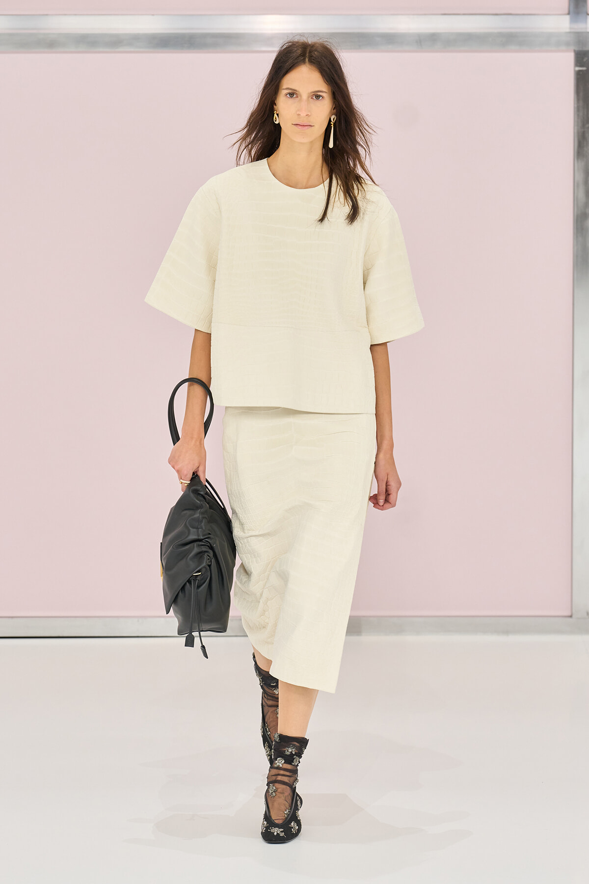 Model in a cream textured two-piece outfit on a runway, carrying a black handbag, with gold earrings and black embellished socks with heels.