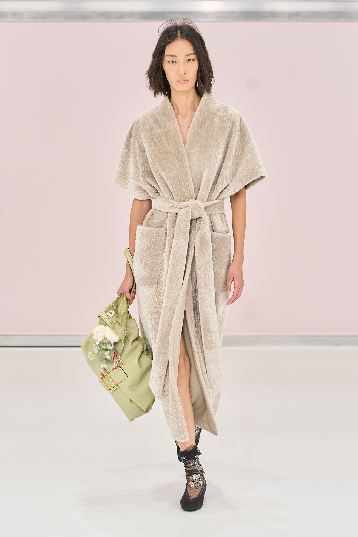 Model walks the runway in a beige plush robe with a belt, carrying a pale green bag.