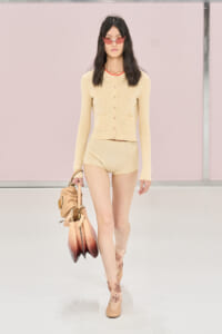 Model walking runway in a pale beige cardigan and matching shorts, red sunglasses, and tan ankle boots, carrying a gradient brown handbag.