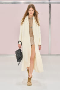 Female model walking a fashion runway in a long cream coat over a beige top and lace shorts, carrying a black handbag.