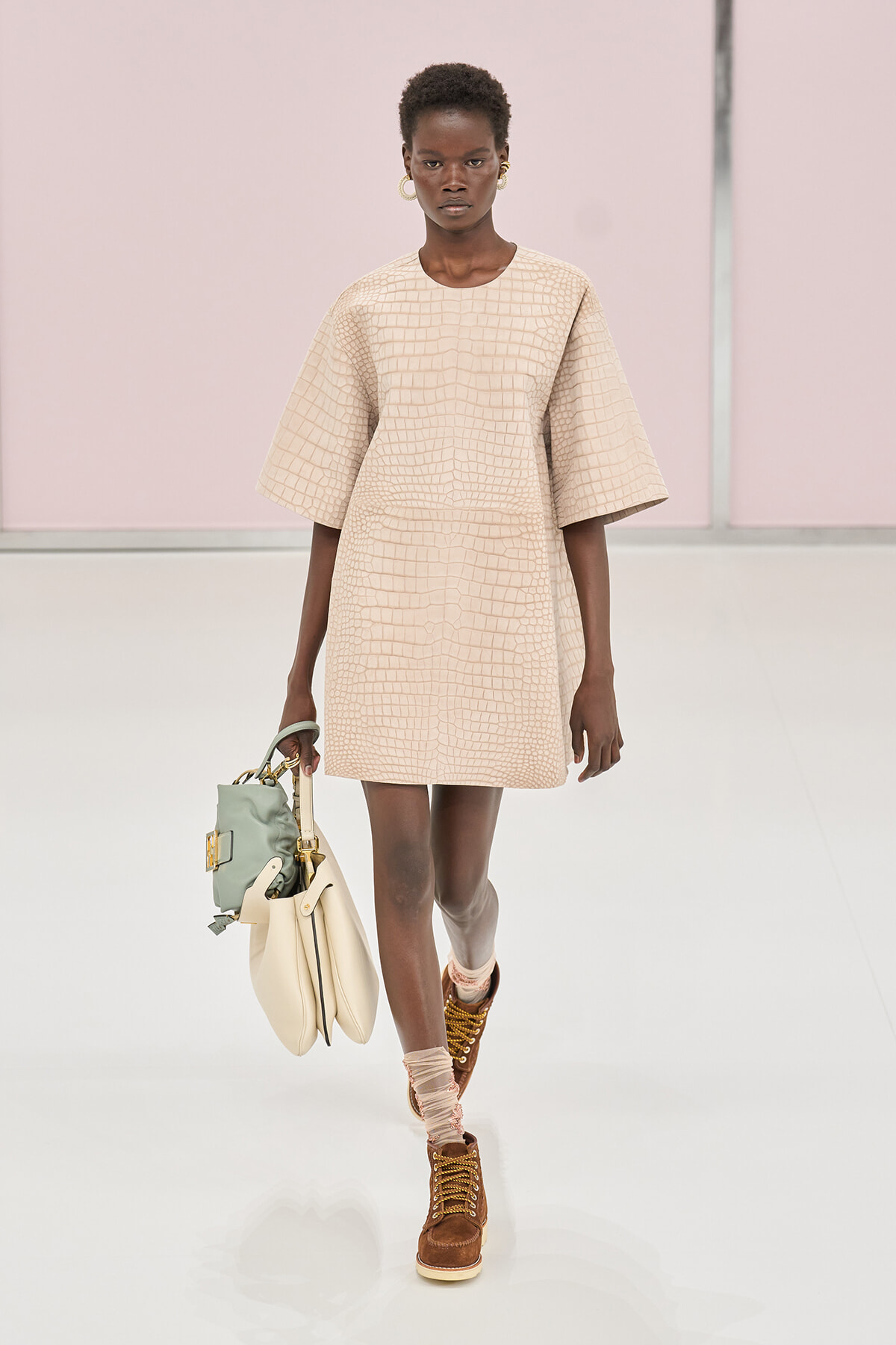 Model walks the runway in a beige crocodile-textured shift dress with oversized sleeves, carrying light-toned bags and wearing brown lace-up boots about mid-calf height.
