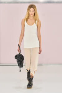 Model walking a runway in a white tank top, sheer cream pants, black combat boots, and a small black handbag.