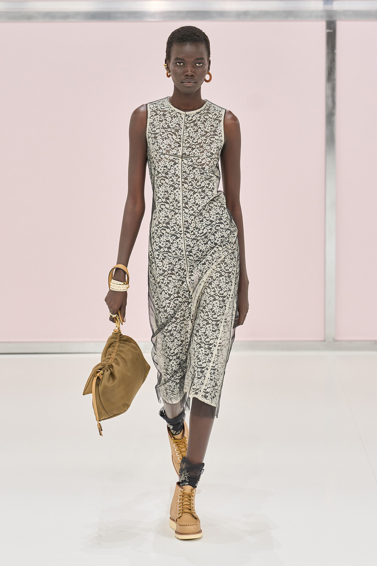 Model walks a runway in a sleeveless ivory lace dress with sheer panels, tan combat boots, and a tan slouch bag.