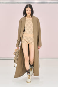 Model walking a runway in a beige floral bodysuit with matching shorts, a long taupe coat, and lace-up boots, carrying a beige handbag.
