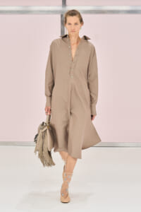 Model walks the runway in a taupe jumpsuit with a button-front placket, carrying a fringed handbag.