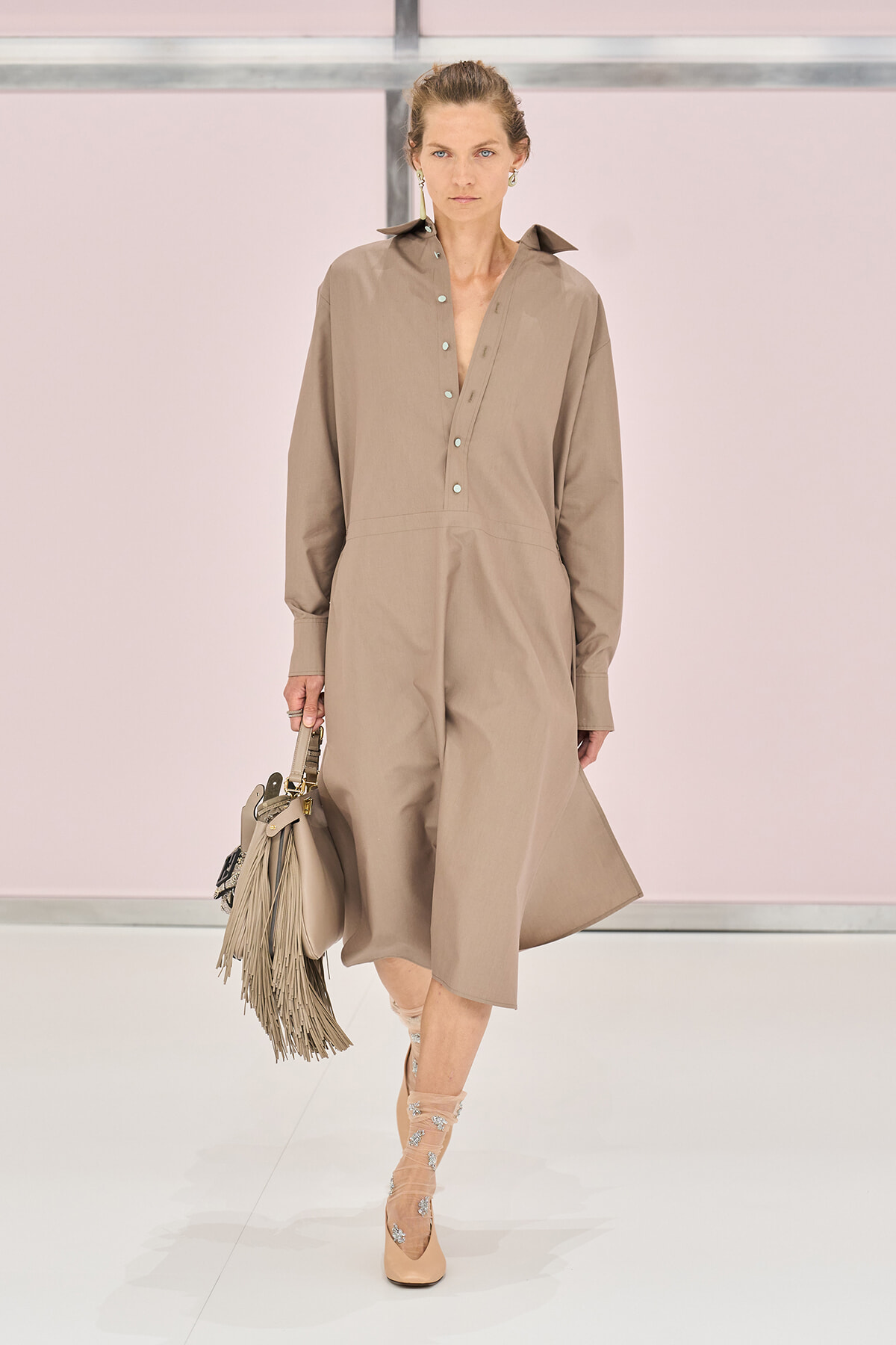Model walks the runway in a taupe jumpsuit with a button-front placket, carrying a fringed handbag.