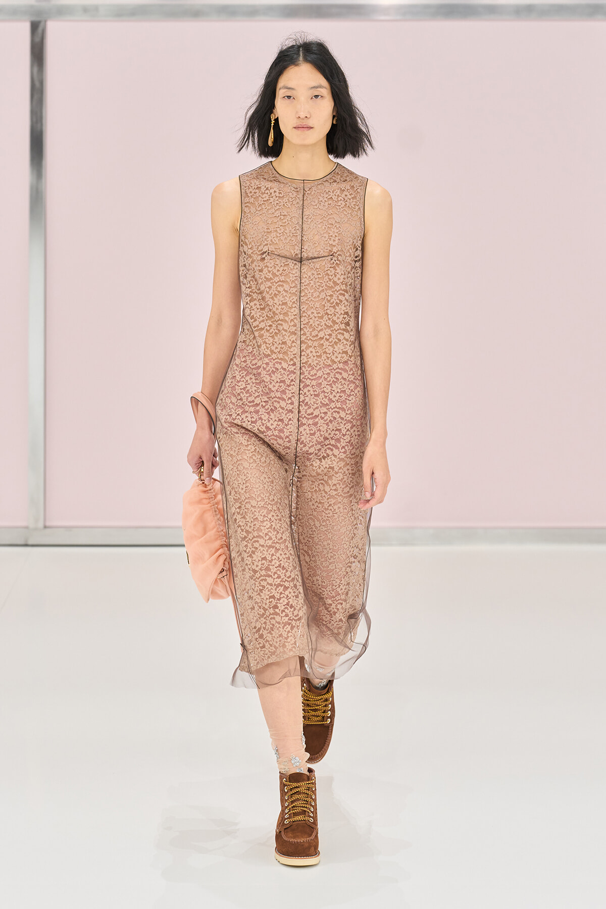 Model walking a runway in a sleeveless nude-pink lace jumpsuit with sheer hem, carrying a peach clutch and wearing brown lace-up boots