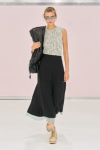 Female model walks the runway in a sleeveless checkered top, wide black trousers with a pale mint hem, beige boots, sunglasses, and a black jacket draped over her shoulder.