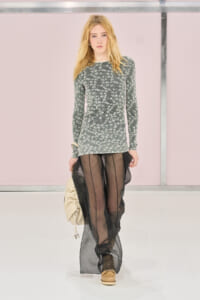 Runway model in a gray floral long-sleeve dress with sheer black pants, tan shoes, and a beige bag against a pink backdrop