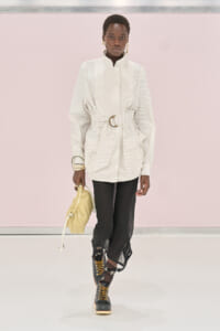 Model walking runway in oversized striped white shirt with wide belt, black sheer pants, and combat boots, carrying a yellow bag and wearing gold jewelry.