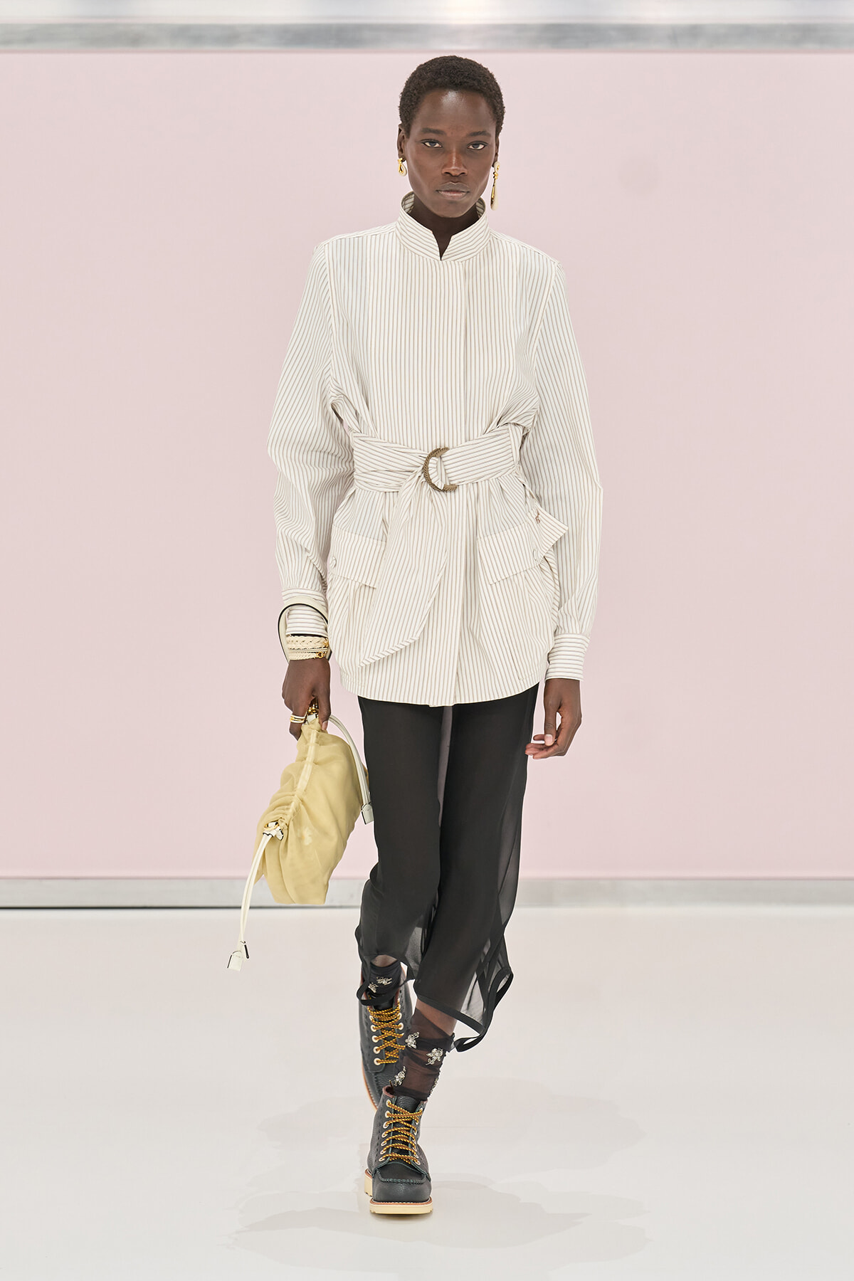 Model walking runway in oversized striped white shirt with wide belt, black sheer pants, and combat boots, carrying a yellow bag and wearing gold jewelry.