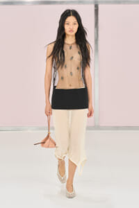 Model walking a runway in a sheer top with embroidered appliqués, a black waist panel, cream cropped pants, white woven heels, and a pink mini bag.