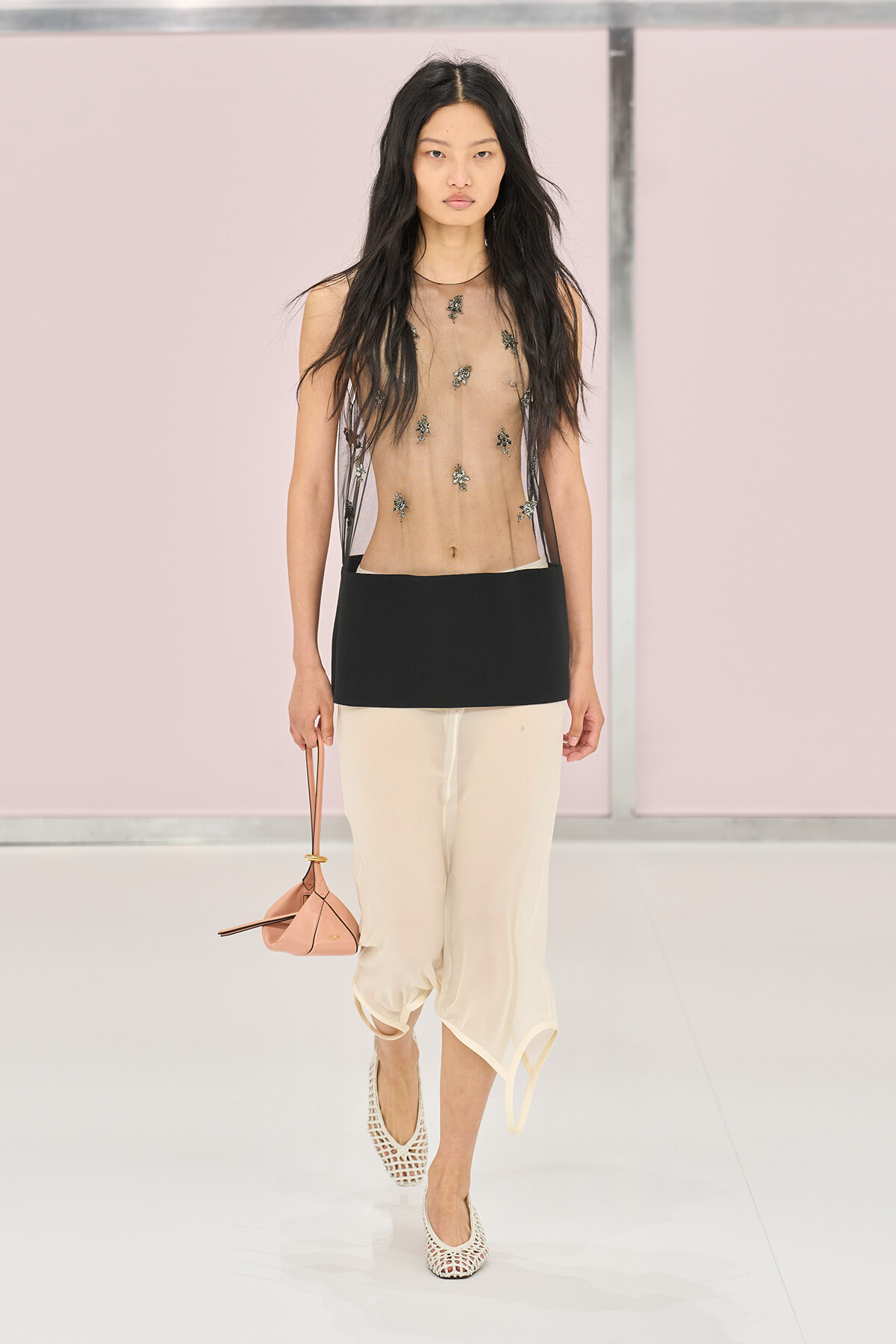 Model walking a runway in a sheer top with embroidered appliqués, a black waist panel, cream cropped pants, white woven heels, and a pink mini bag.