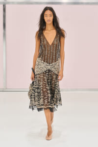 Runway model wearing a sheer brown and black lace dress with a floral-hem pattern and pale pink heels.