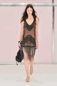 Runway model wearing a sheer black beaded dress with geometric trim and long fringe at the hem.
