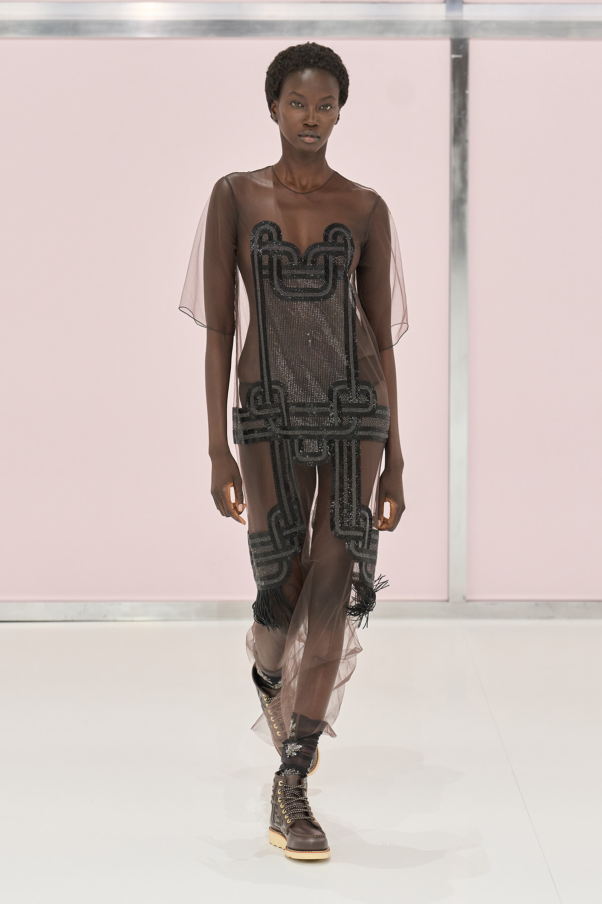 Model on a runway wearing a sheer brown dress with black beaded geometric panels and fringe, paired with lace-up combat boots.