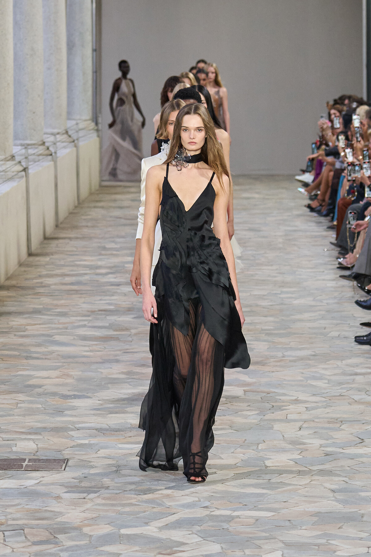 Model walks a fashion runway wearing a black, layered dress with sheer panels and a choker; audience lines the catwalk on both sides.