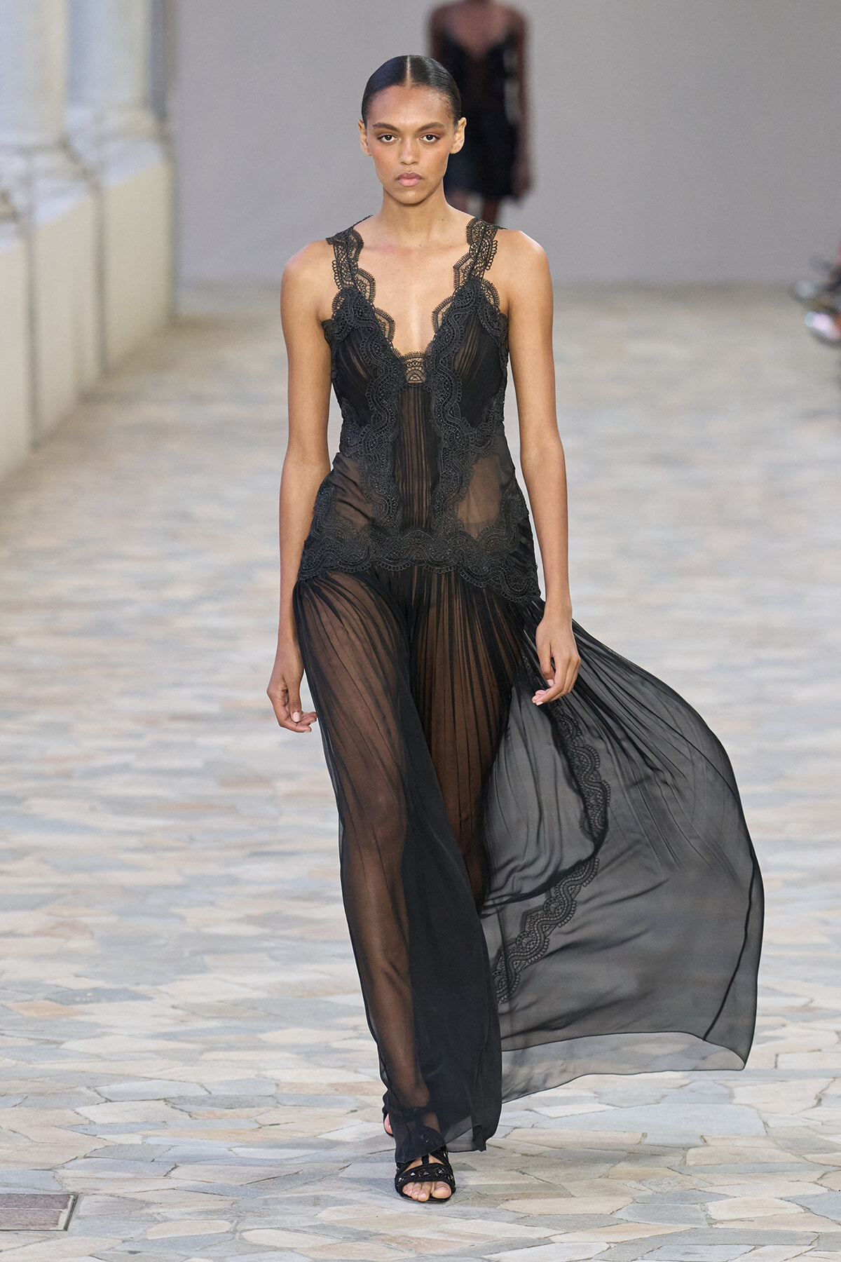 Model walks the runway in a black sheer lace gown with intricate scalloped edging and a flowing, translucent skirt.