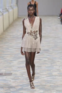 Model walking a beige pleated mini dress with white lace floral appliqué at the bodice and waist, on a stone runway with strappy sandals.