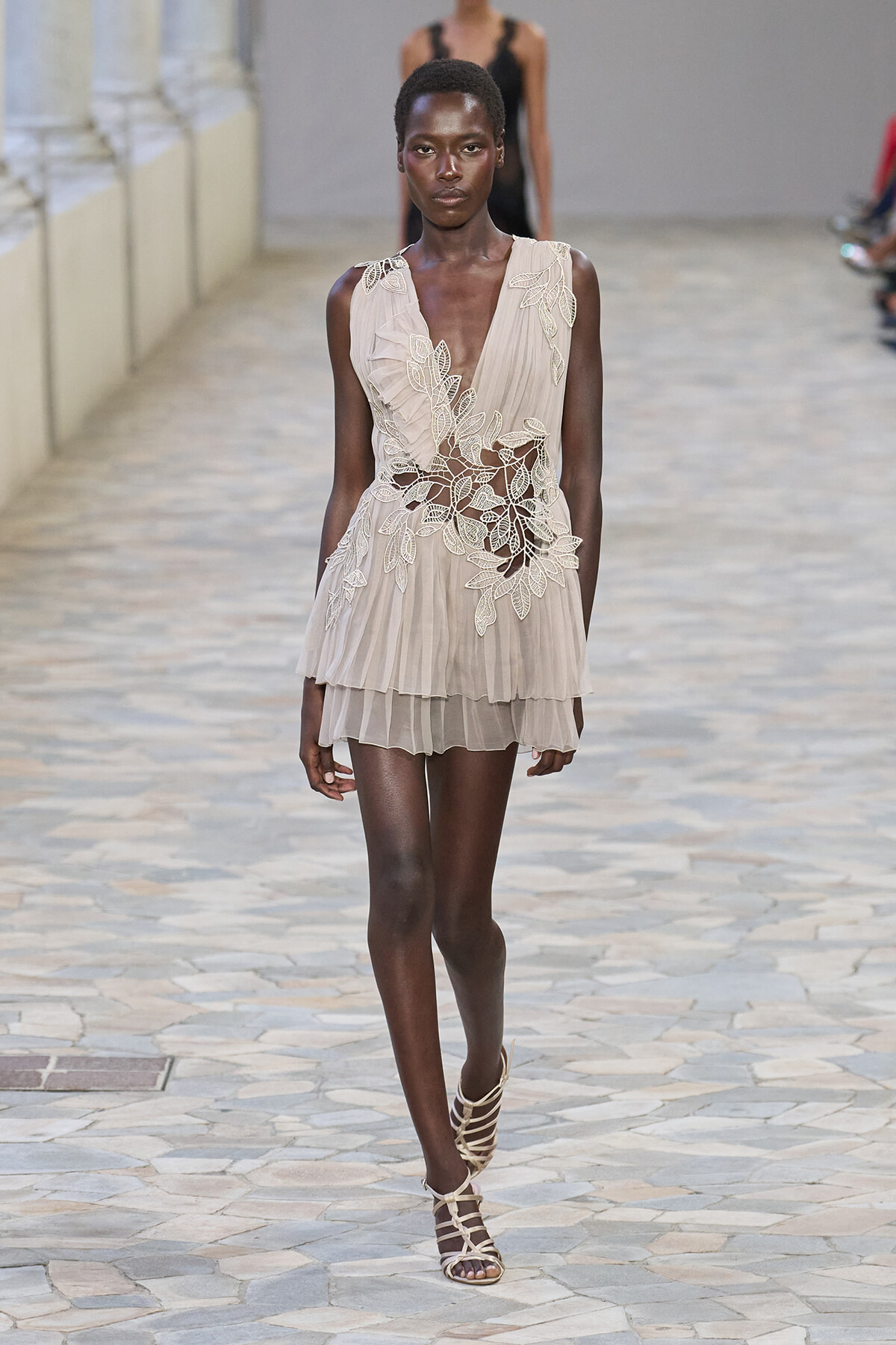 Model walking a beige pleated mini dress with white lace floral appliqué at the bodice and waist, on a stone runway with strappy sandals.