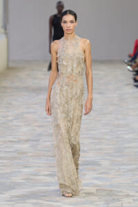 Full-length view of a model walking a runway in a beige, leaf-embellished gown with a high neckline and sheer lace details.