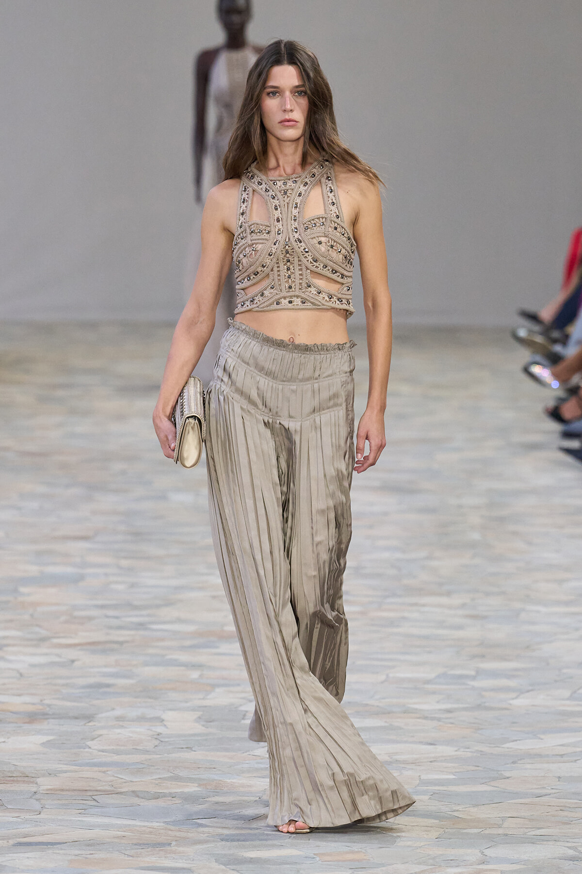 Model walking a runway in a beige beaded two-tone crop top with cutouts and wide metallic-pleated pants, holding a small clutch.