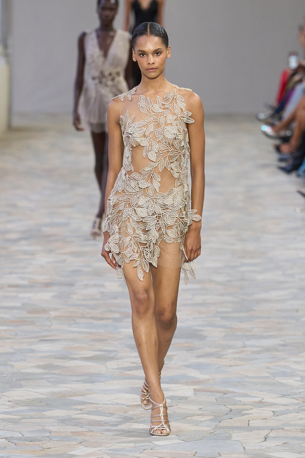 Model walks the runway in a sheer nude dress covered with white leaf lace appliqués.