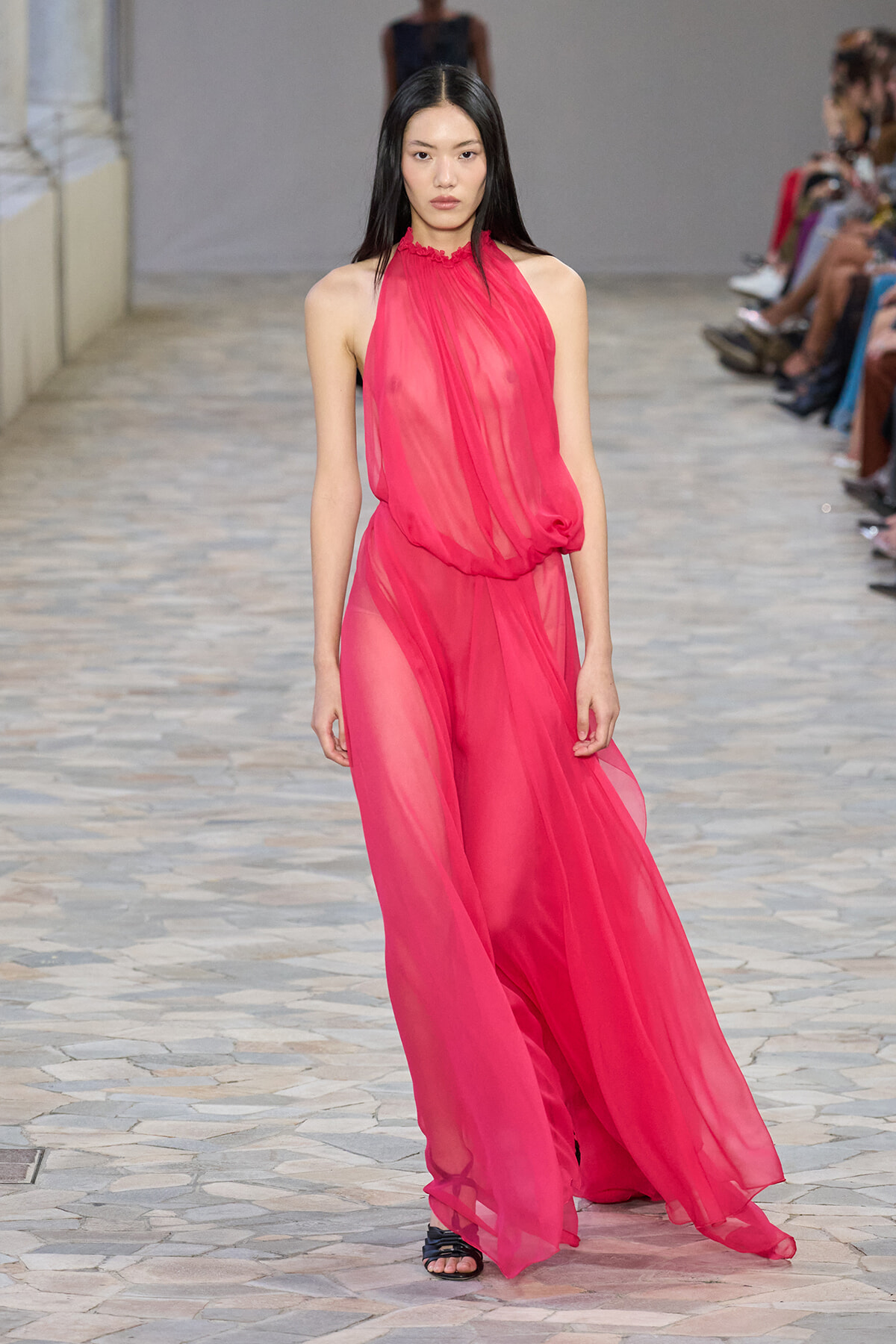 Model walking the runway in a bright pink sheer halter dress with black sandals, on a stone-tiled catwalk.
