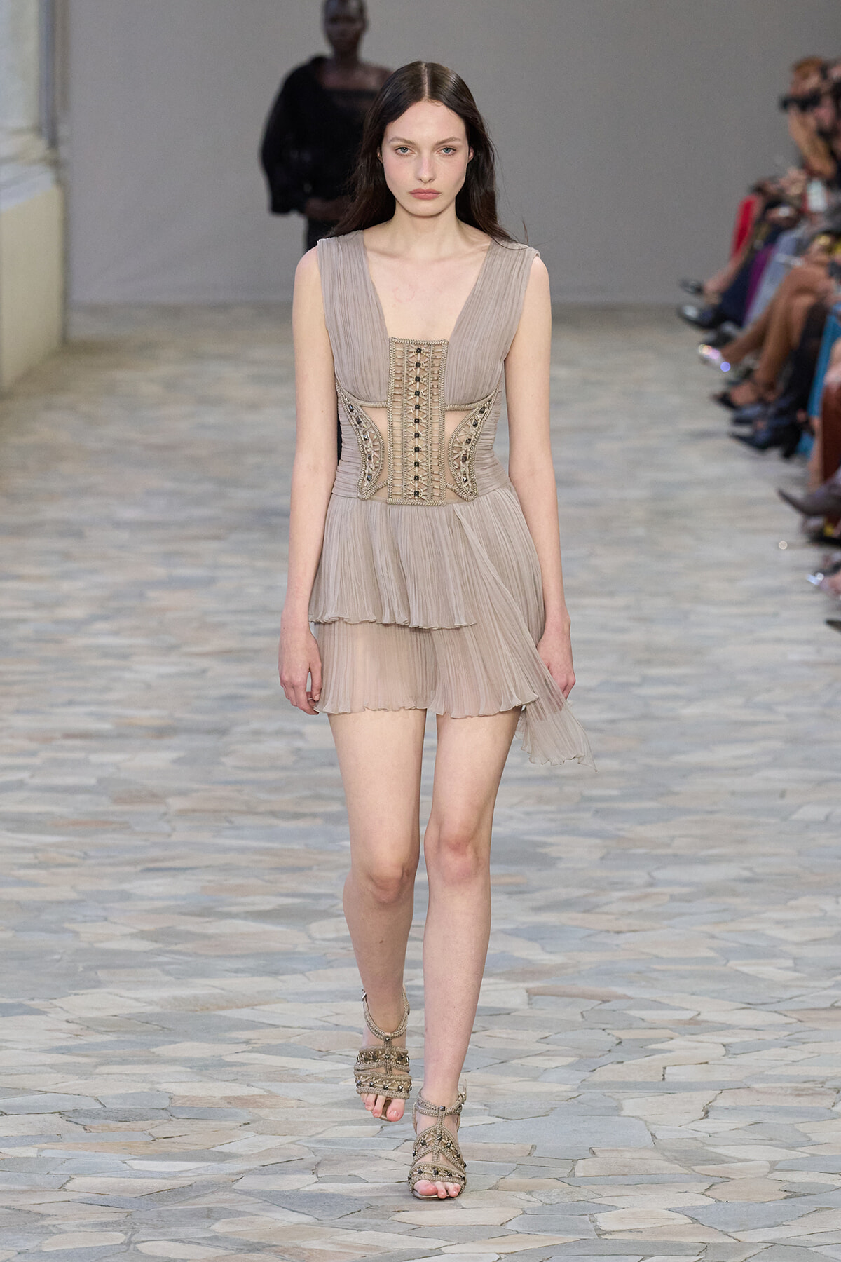 Runway model in a beige, pleated mini dress with an ornate corset-style bodice and strappy sandals.