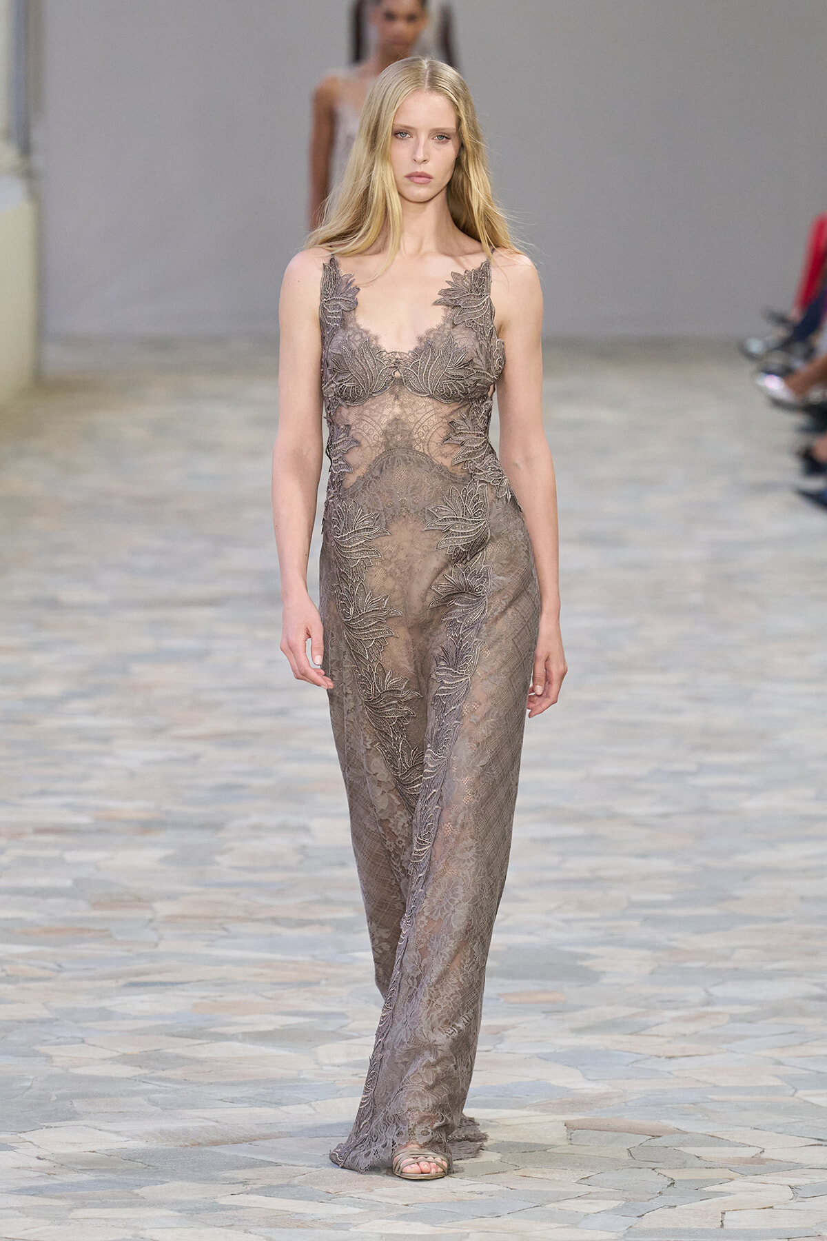 Model walking the runway in a taupe sheer lace gown with leaf appliqué and thin straps.