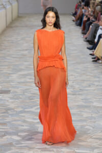 Model walking in a sleeveless orange pleated maxi dress on a tiled runway.