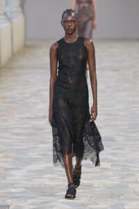 Model on a fashion runway wearing a sleeveless black lace sheer midi dress with scalloped edges and black strappy sandals.