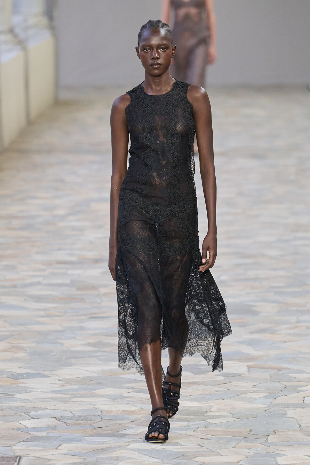 Model on a fashion runway wearing a sleeveless black lace sheer midi dress with scalloped edges and black strappy sandals.