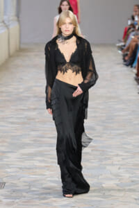 Model walking a runway in a black lace bralette, sheer black blouse, and layered satin skirt with ruffled panels.