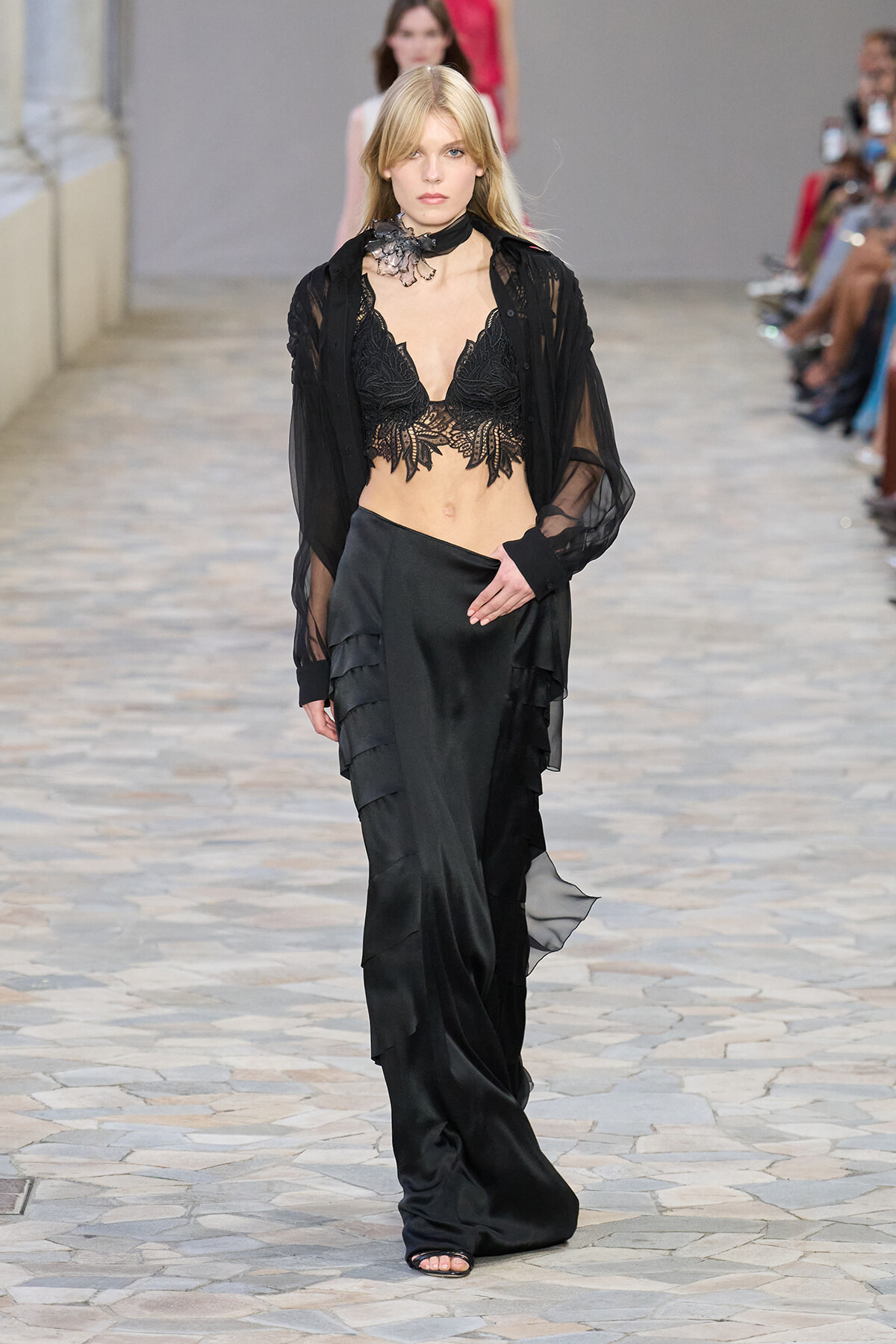 Model walking a runway in a black lace bralette, sheer black blouse, and layered satin skirt with ruffled panels.