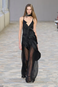 Model walking a stone-paved runway in a black sheer gown with a plunging neckline, ruffled bodice, and long opaque-to-sheer skirt, paired with strappy sandals and a black choker.