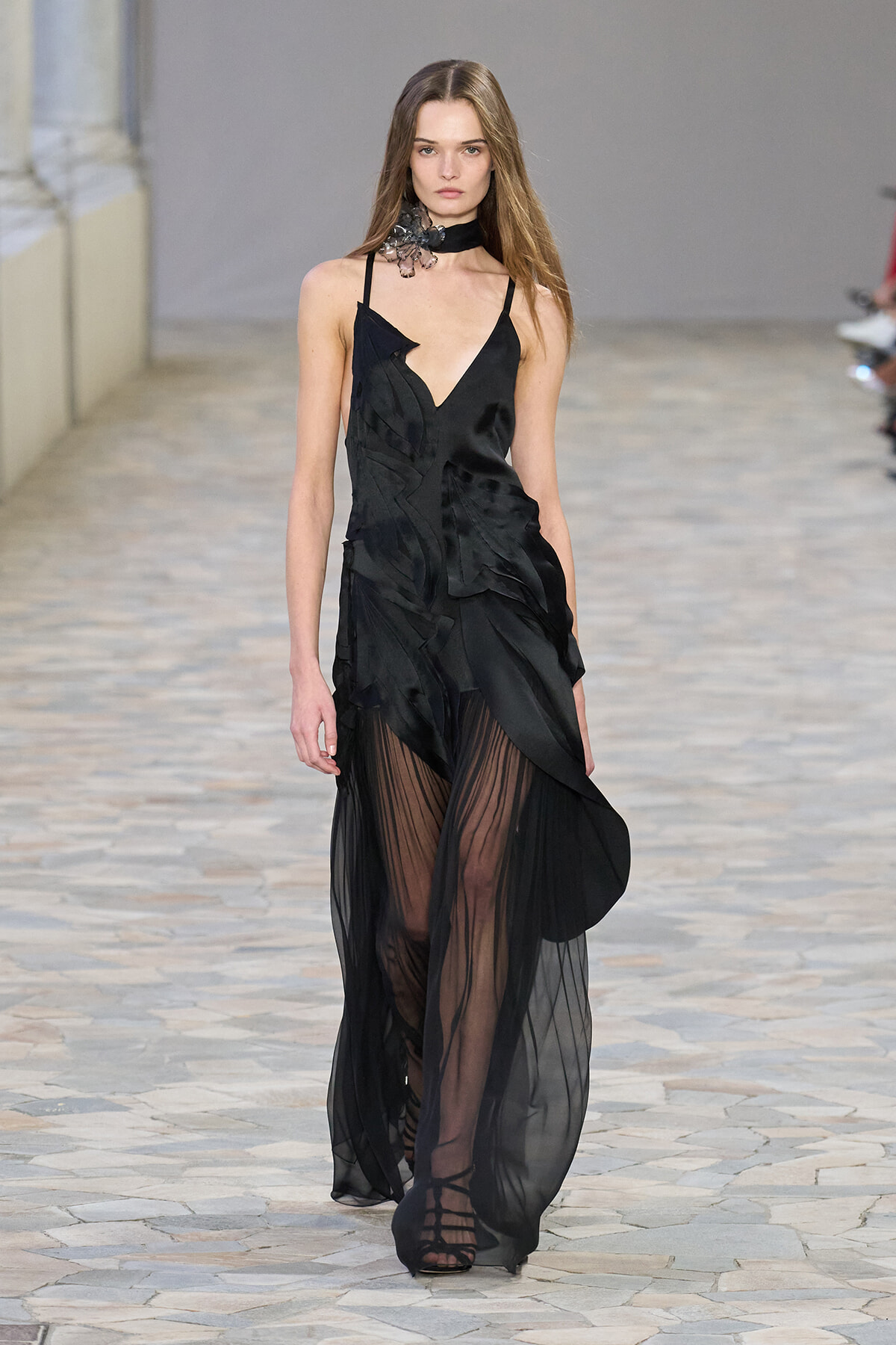 Model walking a stone-paved runway in a black sheer gown with a plunging neckline, ruffled bodice, and long opaque-to-sheer skirt, paired with strappy sandals and a black choker.
