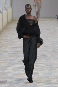 Model walking the runway in an all-black ensemble: off-shoulder, pleated sheer top over a lace bustier with tailored wide-leg pants.