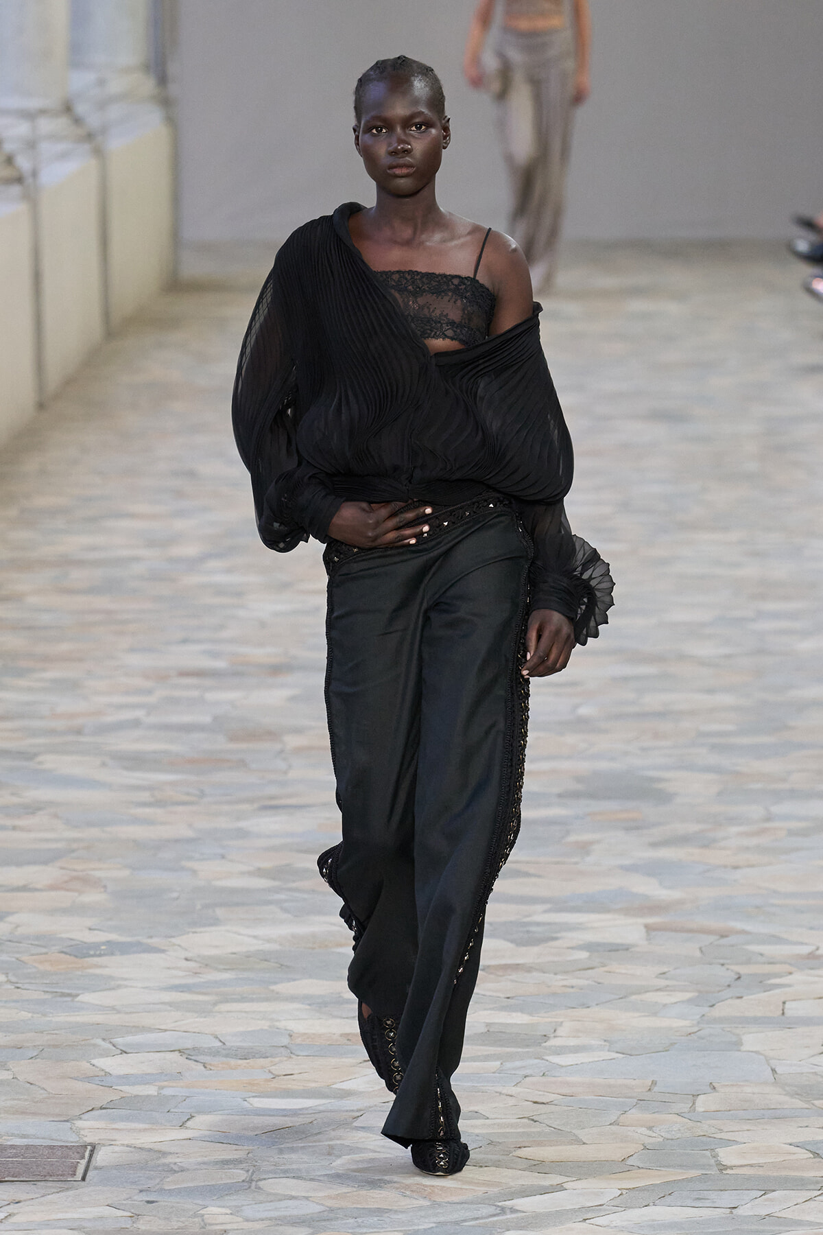Model walking the runway in an all-black ensemble: off-shoulder, pleated sheer top over a lace bustier with tailored wide-leg pants.