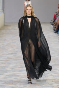 Runway model walking in a sheer black gown with cape-like sleeves and a plunging neckline at a fashion show.